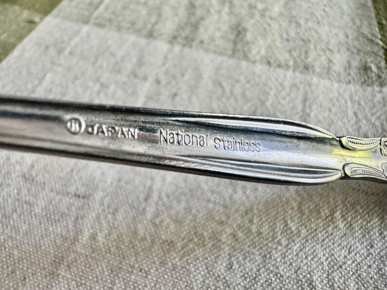 Może przedstawiać: Srebrzysty uchwyt sztućca ze stali nierdzewnej z wytłoczonym wzorem oraz wyraźnie wybitym napisem JAPAN i National stainless na powierzchni. Metal ma polerowane wykończenie z ozdobnymi żłobionymi liniami wzdłuż krawędzi.
