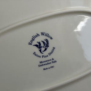 May include: A white ceramic plate base featuring a blue printed logo that reads English Willow Eaton Fine Dining, with an illustration of two birds. The text also states Microwave & Dishwasher Safe and Made in PRC in dark blue lettering.
