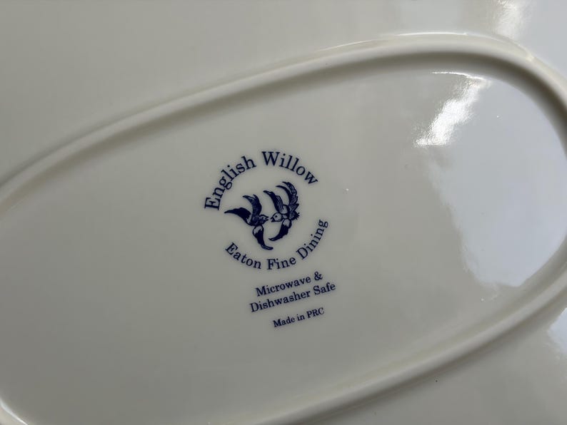 May include: A white ceramic plate base featuring a blue printed logo that reads English Willow Eaton Fine Dining, with an illustration of two birds. The text also states Microwave & Dishwasher Safe and Made in PRC in dark blue lettering.