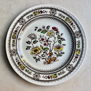 May include: A round ceramic plate featuring a central floral motif with yellow, red, and green accents on a white background. The rim is decorated with a repeating brown and black geometric border interspersed with small floral patterns and scalloped edges.