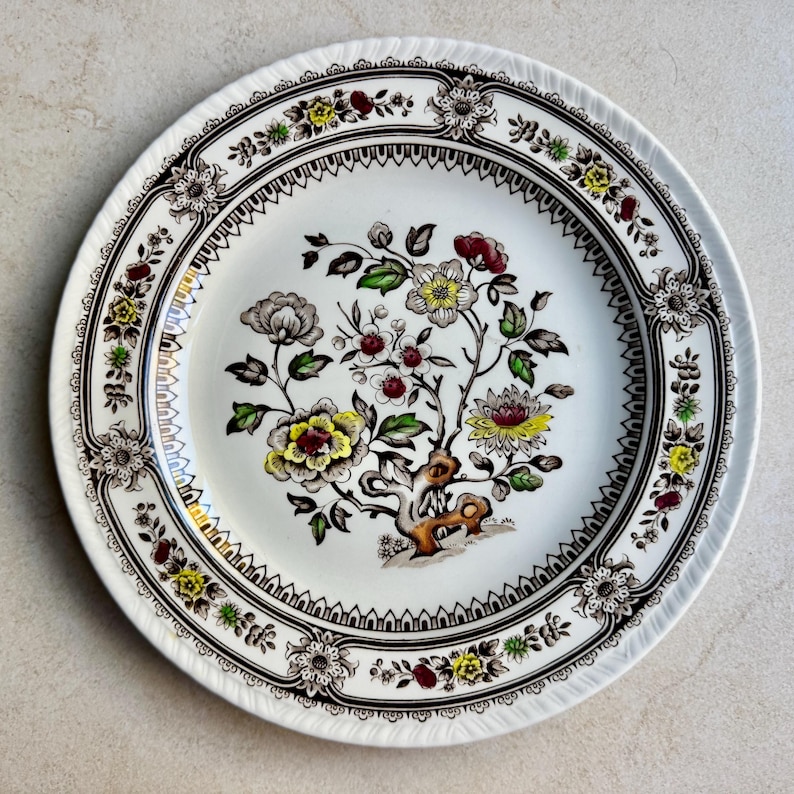 May include: A round ceramic plate featuring a central floral motif with yellow, red, and green accents on a white background. The rim is decorated with a repeating brown and black geometric border interspersed with small floral patterns and scalloped edges.