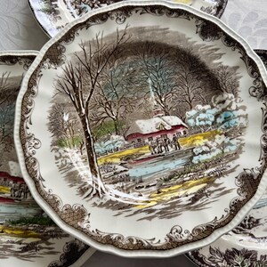 May include: Set of four vintage ceramic dinner plates featuring a winter scene with a snow-covered house, trees, and a frozen pond. The plates have a brown and white color scheme with intricate floral details around the rim.