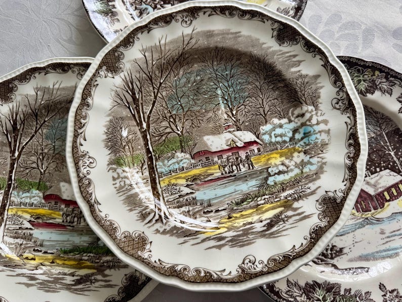 May include: Set of four vintage ceramic dinner plates featuring a winter scene with a snow-covered house, trees, and a frozen pond. The plates have a brown and white color scheme with intricate floral details around the rim.