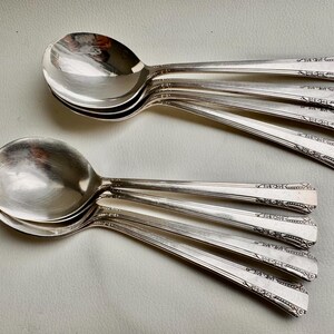 May include: A set of eight silver soup spoons with ornate handles. The spoons are stacked on top of each other, with the handles extending out to the sides.