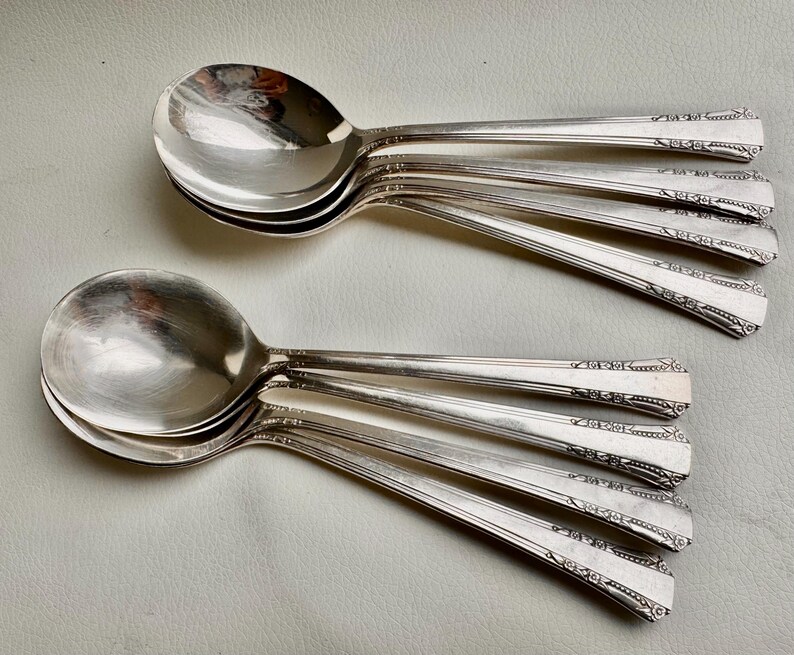 May include: A set of eight silver soup spoons with ornate handles. The spoons are stacked on top of each other, with the handles extending out to the sides.