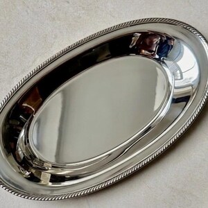 K&ouml;nnte beinhalten: Ein ovales Serviertablett in Silberoptik mit einer polierten, reflektierenden Oberfl&auml;che. Das Tablett verf&uuml;gt &uuml;ber einen erhabenen, strukturierten Rand im Seil-Design, der eine klare Abgrenzung zur glatten, konkaven Mitte des Tabletts bildet.