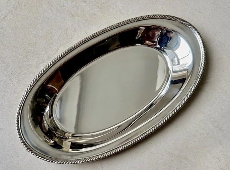 K&ouml;nnte beinhalten: Ein ovales Serviertablett in Silberoptik mit einer polierten, reflektierenden Oberfl&auml;che. Das Tablett verf&uuml;gt &uuml;ber einen erhabenen, strukturierten Rand im Seil-Design, der eine klare Abgrenzung zur glatten, konkaven Mitte des Tabletts bildet.