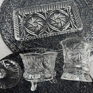 May include: A clear pressed glass set consisting of a rectangular tray, a lidded sugar bowl with three feet, and a creamer pitcher. Each piece features a detailed starburst and geometric pattern with scalloped edges and a brilliant reflective finish.
