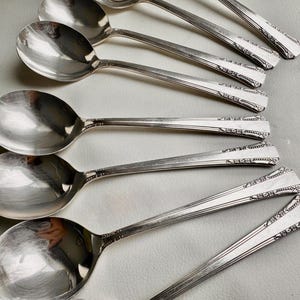 May include: A set of eight silver spoons with ornate designs on the handles. The spoons are arranged in a row, with the handles pointing to the right.