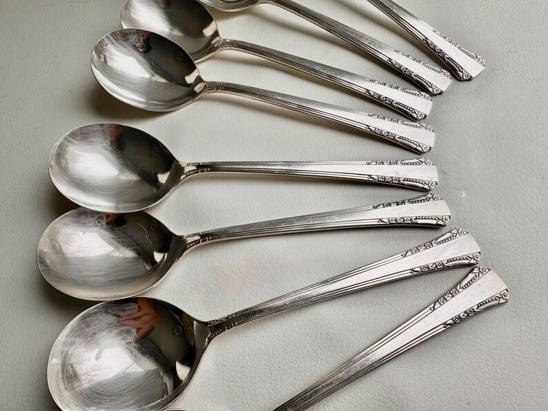 May include: A set of eight silver spoons with ornate designs on the handles. The spoons are arranged in a row, with the handles pointing to the right.