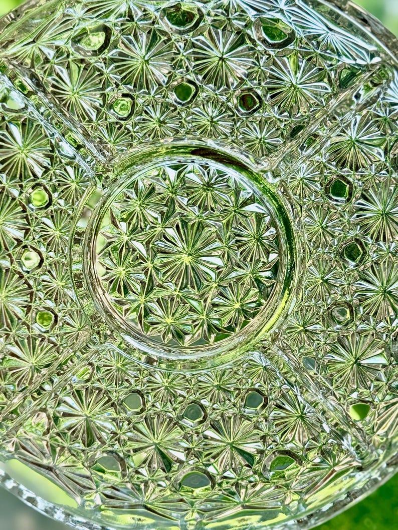 May include: Close-up of a clear, vintage glass serving dish with a starburst pattern. The dish is divided into sections and has a circular center. The intricate design reflects light, creating a sparkling effect. The dish is likely used for serving snacks or appetizers.