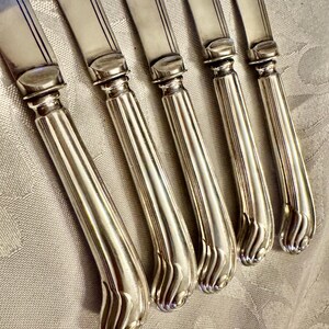 K&ouml;nnte beinhalten: Nahaufnahme von sechs silbernen Messern. Die Messer haben ein klassisches Design mit einer gl&auml;nzenden, reflektierenden Oberfl&auml;che. Die Griffe sind gebogen und mit einem dekorativen, geriffelten Muster versehen. Die Klingen sind glatt und rechteckig. Die Messer sind in einer Reihe angeordnet, leicht abgewinkelt.