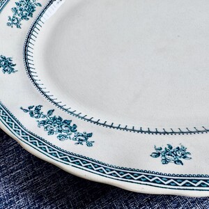 May include: An oval ceramic serving platter with an off-white base colour. The rim features a dark teal geometric border pattern and a series of small, repeating floral clusters in the same dark teal colour, arranged along the edge.