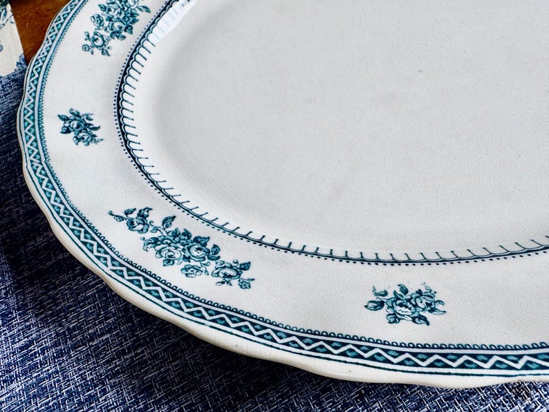 May include: An oval ceramic serving platter with an off-white base colour. The rim features a dark teal geometric border pattern and a series of small, repeating floral clusters in the same dark teal colour, arranged along the edge.