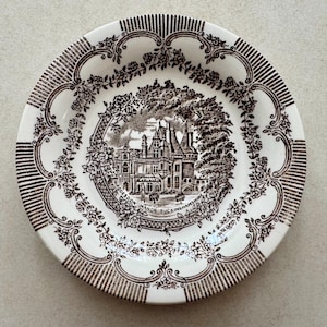 May include: A round ceramic plate featuring a dark brown transferware pattern on a cream background. The center depicts Waddesdon Manor Buckinghamshire surrounded by a floral wreath. The rim has a decorative border of roses and vertical lines.