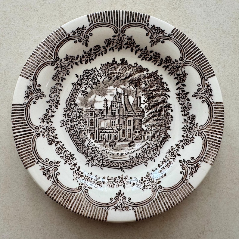 May include: A round ceramic plate featuring a dark brown transferware pattern on a cream background. The center depicts Waddesdon Manor Buckinghamshire surrounded by a floral wreath. The rim has a decorative border of roses and vertical lines.