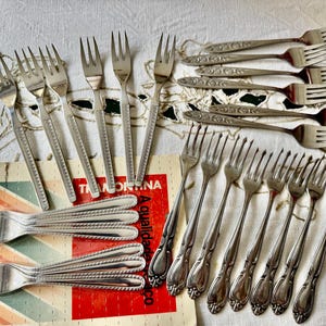 May include: An assortment of vintage silver forks, some with ornate floral designs and others with simple, elegant lines. The forks are arranged on a patterned tablecloth and a Tramontina box.