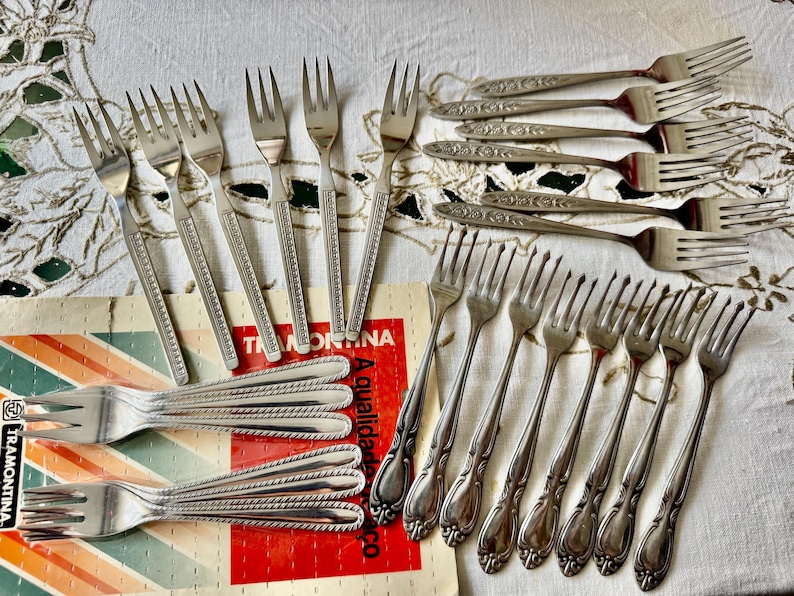 May include: An assortment of vintage silver forks, some with ornate floral designs and others with simple, elegant lines. The forks are arranged on a patterned tablecloth and a Tramontina box.