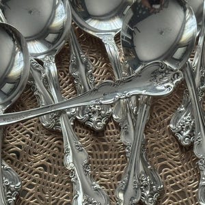 May include: A collection of ornate silver spoons and ladles. The silverware features detailed floral and scrollwork patterns. The spoons and ladles are arranged on a textured, golden-brown surface, reflecting light.