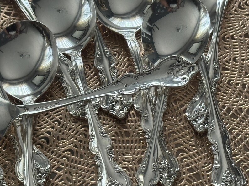 May include: A collection of ornate silver spoons and ladles. The silverware features detailed floral and scrollwork patterns. The spoons and ladles are arranged on a textured, golden-brown surface, reflecting light.