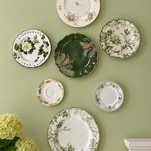 May include: A collection of six decorative ceramic plates arranged on a wall. The plates feature various designs including floral patterns, a dark green plate with pinecone and needle motifs, and intricate green and white botanical illustrations.