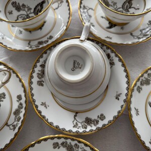 May include: Set of five white teacups and saucers with a black and gold floral pattern. The saucers have a gold rim and the teacups have a gold band around the top. The teacups have a gold inscription on the bottom that reads "Crescent"