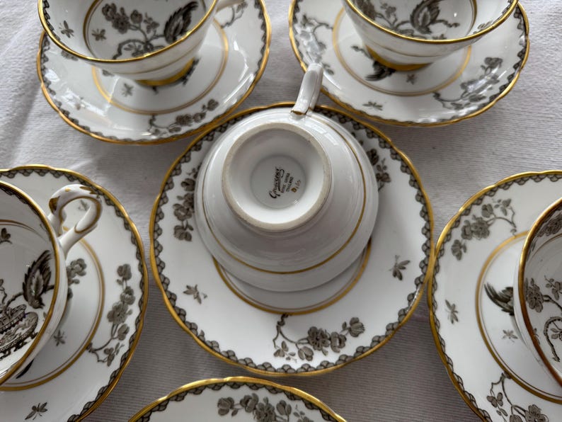 May include: Set of five white teacups and saucers with a black and gold floral pattern. The saucers have a gold rim and the teacups have a gold band around the top. The teacups have a gold inscription on the bottom that reads "Crescent"