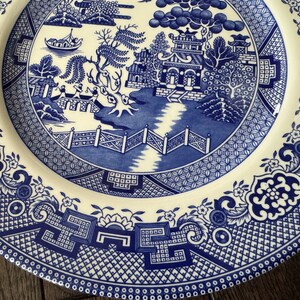 K&ouml;nnte beinhalten: Ein blauer und wei&szlig;er Keramikteller mit einem traditionellen chinesischen Weidenmuster. Der Teller zeigt ein zentrales Design mit einer Pagode, B&auml;umen und einer Br&uuml;cke, umgeben von einem Rand aus sich wiederholenden floralen und geometrischen Motiven.