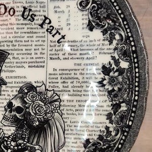K&ouml;nnte beinhalten: Ein schwarzer und wei&szlig;er Keramikteller mit einem dekorativen Rand und einer Illustration eines Totenkopfes und einer Skelettbraut und eines Br&auml;utigams. Der Teller zeigt Text &uuml;ber die Gro&szlig;e Ausstellung von 1851.