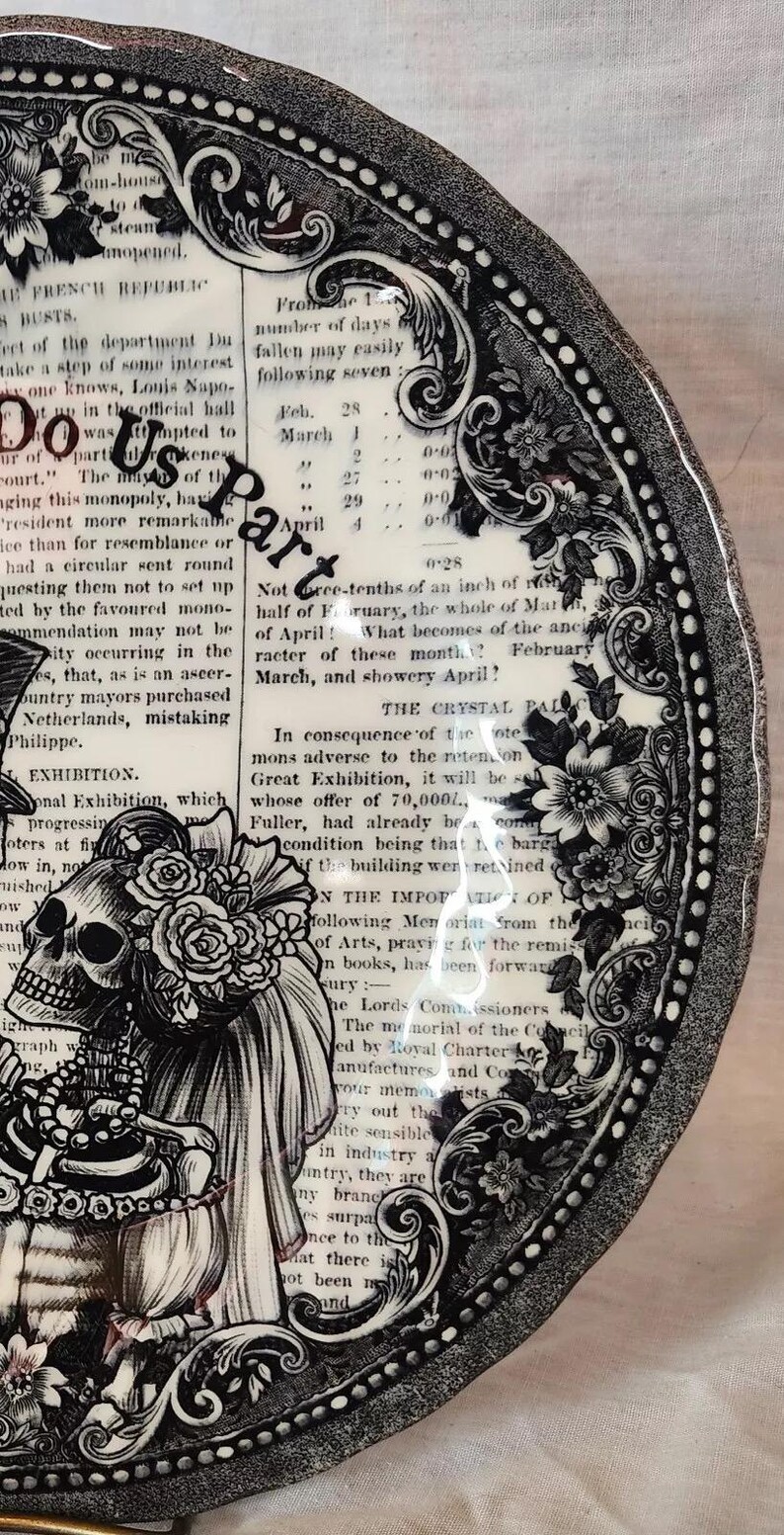 K&ouml;nnte beinhalten: Ein schwarzer und wei&szlig;er Keramikteller mit einem dekorativen Rand und einer Illustration eines Totenkopfes und einer Skelettbraut und eines Br&auml;utigams. Der Teller zeigt Text &uuml;ber die Gro&szlig;e Ausstellung von 1851.