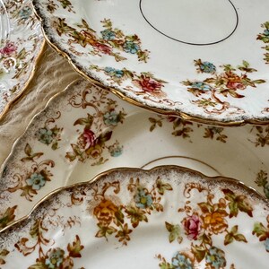 K&ouml;nnte beinhalten: Ein Satz von vier Vintage-Tellern mit floralem Muster und goldener Verzierung. Die Teller sind wei&szlig; mit einem zarten Blumenmuster in Rosa-, Blau- und Gelbt&ouml;nen. Die Teller sind &uuml;bereinander gestapelt, wobei der oberste Teller leicht versetzt ist.