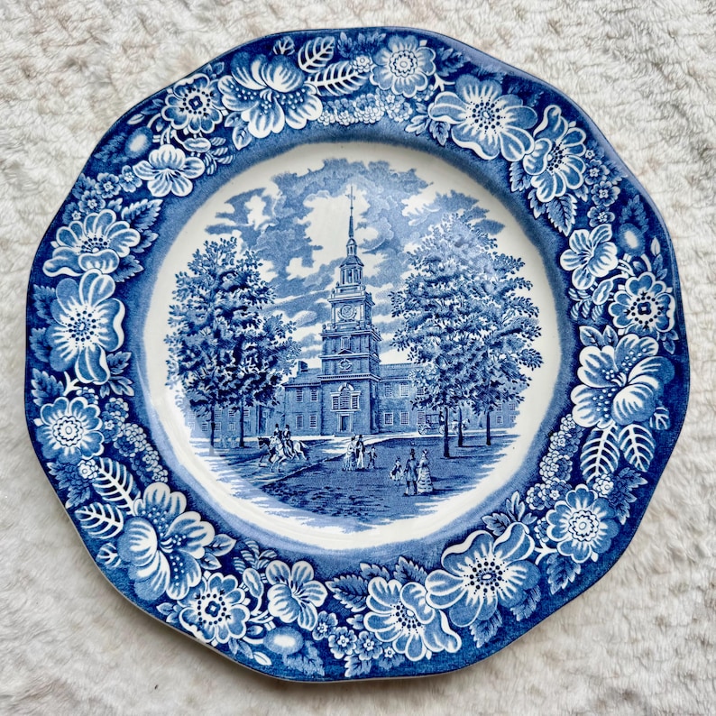 May include: A round ceramic plate featuring a blue and white transferware pattern. The center depicts a historic building with a tall steeple, trees, and figures. The wide rim is decorated with a dense border of various flowers and leaves.