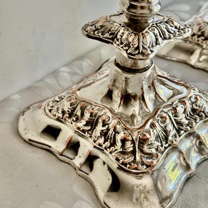 May include: A silver-plated candlestick base featuring an ornate, square-shaped pedestal with intricate embossed floral and scrollwork detailing. The metal surface displays a polished finish with visible copper-toned wear along the edges.