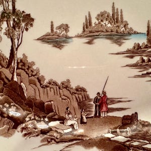 Op de afbeelding: Een keramieken bord met een bruine transferprint van een landschap. De afbeelding toont een rivier met een waterrad, bomen, rotsachtig terrein en figuren die aan de oever staan. De sc&egrave;ne bevat lichtblauwe accenten op het water.