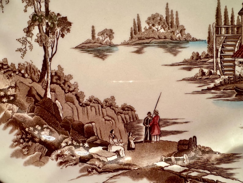 Op de afbeelding: Een keramieken bord met een bruine transferprint van een landschap. De afbeelding toont een rivier met een waterrad, bomen, rotsachtig terrein en figuren die aan de oever staan. De sc&egrave;ne bevat lichtblauwe accenten op het water.