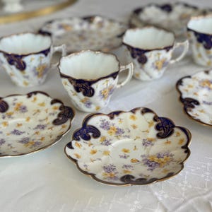 May include: A set of vintage porcelain teacups and saucers. The cups and saucers have a white base with a floral design in yellow and purple. The rims and handles are trimmed with gold and dark blue details. The set is displayed on a white tablecloth.