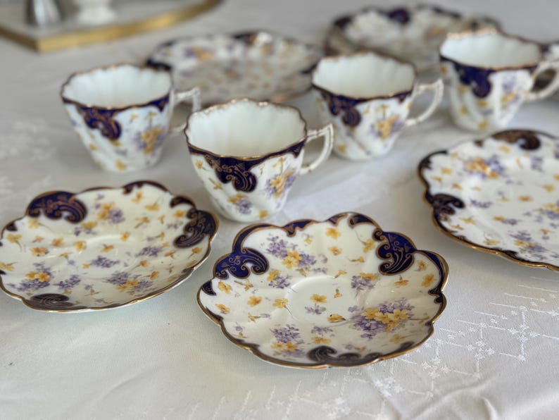 May include: A set of vintage porcelain teacups and saucers. The cups and saucers have a white base with a floral design in yellow and purple. The rims and handles are trimmed with gold and dark blue details. The set is displayed on a white tablecloth.