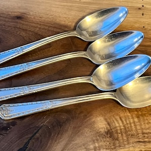 Pu&ograve; includere: Quattro cucchiai d'argento con dettagli floreali ornati sui manici e una ciotola ovale riflettente. I cucchiai sono disposti diagonalmente su una superficie di legno scuro, mettendo in risalto la loro finitura lucida.
