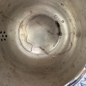 May include: A silver-toned metal colander with a circular basin featuring a series of small drainage holes on the side. The interior surface shows visible water droplets and faint dark residue marks across the bottom of the bowl.