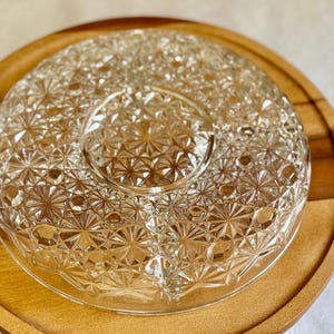 May include: A clear, starburst-patterned glass serving dish with a central dip section, set on a round wooden tray with a handle. The dish is designed for serving appetizers or snacks.