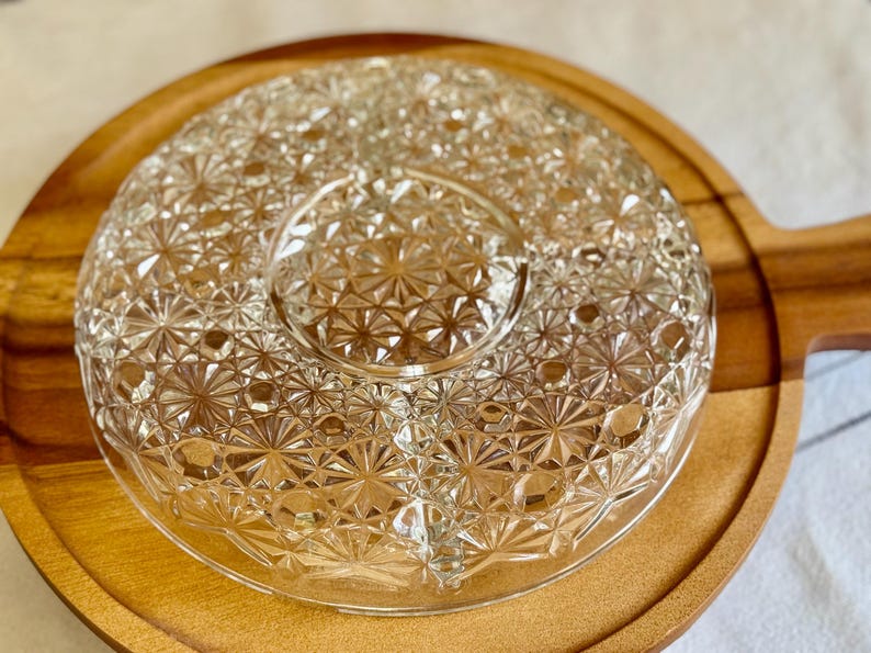 May include: A clear, starburst-patterned glass serving dish with a central dip section, set on a round wooden tray with a handle. The dish is designed for serving appetizers or snacks.