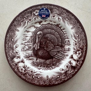 May include: A round ceramic plate featuring a brown and white transferware pattern. The center depicts a turkey in a rural landscape with a house. The rim is decorated with wheat, berries, and flowers. A Royal Stafford logo is at the top.