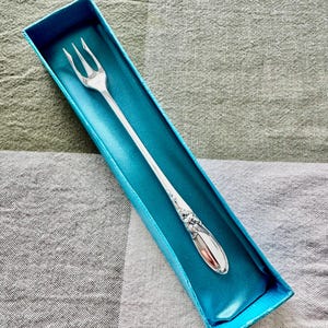 Op de afbeelding: Een zilveren vork met drie tanden, gepresenteerd in een turquoise blauwe doos. De vork heeft een decoratief bloemmotief op het handvat. De doos is gevoerd met een satijnachtig materiaal, tegen een geruite achtergrond.