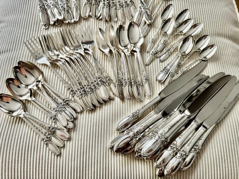 Op de afbeelding: Een assortiment zilverkleurig bestek, waaronder vorken, lepels en messen, gerangschikt op een gestreept oppervlak. Het zilverwerk heeft een bloemmotief op de handvatten. De lepels, vorken en messen zijn in groepen gerangschikt.