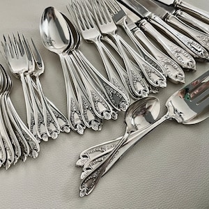 Puede incluir: Un conjunto de cubiertos de plata pulida, que incluye cucharas, tenedores y cuchillos. La cuberter&iacute;a presenta detalles florales ornamentados en los mangos. La disposici&oacute;n incluye utensilios para servir, mostrando un juego completo para cenar.