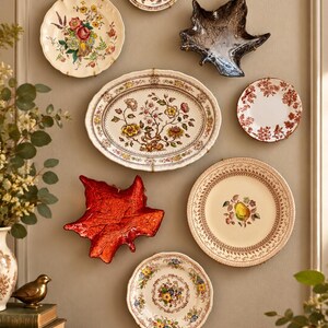 May include: A collection of seven decorative wall plates and two leaf-shaped dishes arranged on a wall. The items feature various floral, fruit, and botanical patterns in colors including red, yellow, green, and brown, alongside a dark grey and a red leaf dish.