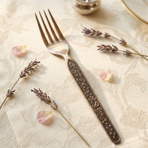 Puede incluir: Un tenedor de metal plateado con un intrincado patr&oacute;n floral en relieve a lo largo del mango. El tenedor est&aacute; colocado en diagonal, acompa&ntilde;ado por varias ramas de lavanda seca y peque&ntilde;os p&eacute;talos de flores rosas sobre una tela color crema.