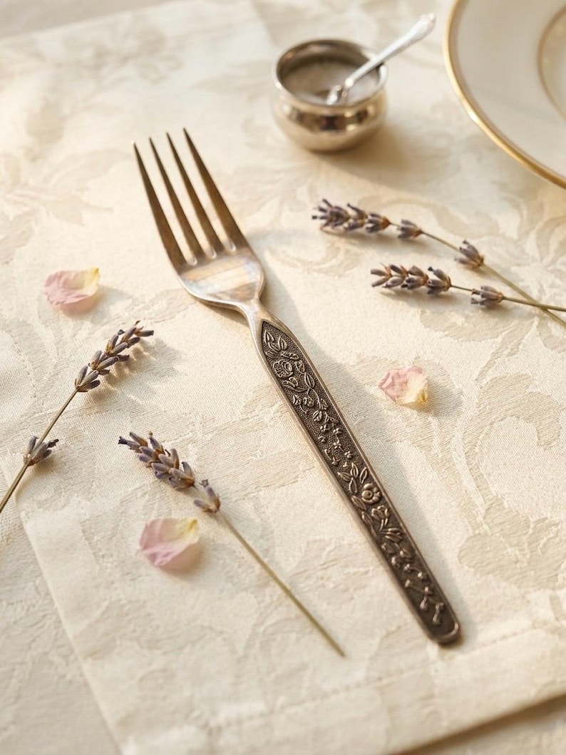 Puede incluir: Un tenedor de metal plateado con un intrincado patr&oacute;n floral en relieve a lo largo del mango. El tenedor est&aacute; colocado en diagonal, acompa&ntilde;ado por varias ramas de lavanda seca y peque&ntilde;os p&eacute;talos de flores rosas sobre una tela color crema.