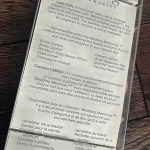 May include: A white card with the text "Wedding Memories" in black lettering. The card lists various wedding-related items, including a cake knife, garters, a keepsake bag, and toasting glasses. The text "Wedding Memories" is a trademark.