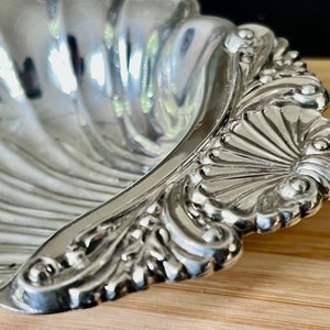 May include: A silver shell-shaped dish with a decorative, ornate rim. The dish is made of polished silver and has a reflective surface.